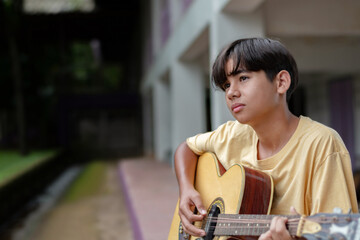 Teen boy playing acoustic guitar outdoors, wearing a yellow shirt, looking thoughtful. Creative youth expression and musical lifestyle in a casual setting.
