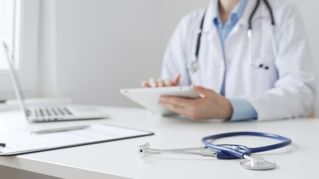 Closeup of blue stethoscope is lying on the table near female doctor reviewing patient data on tablet, representing digital healthcare workflow in contemporary medical setting - Powered by Adobe