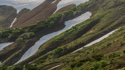 Scenic view of rolling hills with lush green vegetation and patches of snow, showcasing the beauty of nature and the interplay of light and shadow in a tranquil landscape