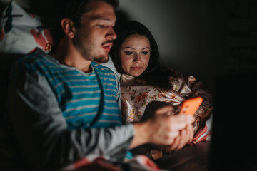 A couple is sitting in bed, illuminated by the smart phone screen, sharing a moment of relaxation. The ambient light creates a cozy, intimate atmosphere as they engage with technology.