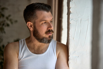 Pensive bearded man wearing a white tank top gazes out a window, bathed in soft natural light...