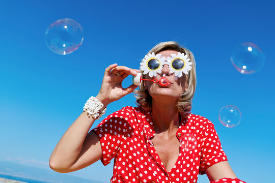 Blond woman in red polka dot dress blowing soap bubbles on a sunny day at the beach