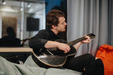 Teen boy artistically playing an acoustic black guitar on a cozy indoor sofa. The scene portrays a...