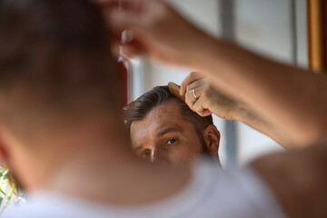 Clean-boy grooming. Caucasian man is styling his hair while looking in the mirror with a focused expression. Concept of men's grooming and personal care routines.