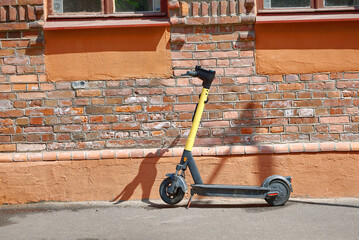 Yellow rental scooter parked on city sidewalk beside vintage red brick building under the warm sunlight on clear summer day in an urban environment. Eco-friendly scooter near historic building