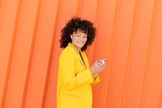 Happy woman wearing yellow blazer listening to music and dancing in front of orange wall