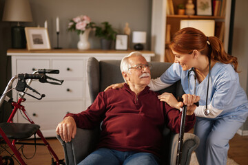 Inside a comfortable home, a kind red-haired nurse in blue scrubs gently rests her hands on an...