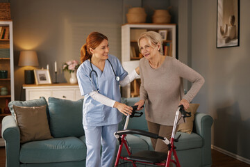 A young, red-haired caregiver in blue scrubs assists an elderly woman with a walker in her living...