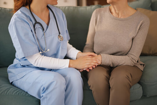A healthcare worker, wearing blue scrubs and a stethoscope, sits beside an older woman on a sofa. She holds the woman's hands in a comforting gesture during a home visit.