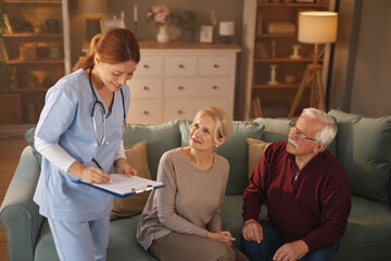 A female nurse in blue scrubs takes notes on a clipboard while talking to an elderly couple who are...