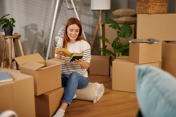 Surrounded by unpacked boxes, a woman sits comfortably, reading a book and enjoying a peaceful...