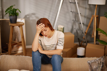 A woman sits on a couch in a partially packed living room, looking stressed and overwhelmed during...