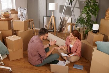 Two individuals are sitting on the floor, engaged in conversation amidst numerous moving boxes in...