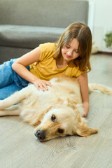 A young girl is lying on a wooden floor, happily interacting with a golden retriever. They are in a light-filled living room, enjoying each other's company and playing together.
