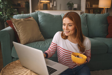 In a warm living room, a woman smiles as she interacts with her laptop while holding a bowl of...