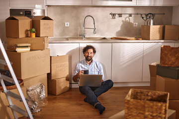 A person sits on the floor surrounded by moving boxes, using a laptop in a kitchen environment...