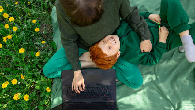 Mother and red-haired son lying on grass working remotely on a laptop
