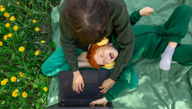 Red-haired boy playfully interrupts his mother working on a laptop outdoors.