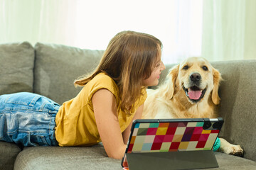 A teen girl lies comfortably on the couch in a cozy living room, engaging with her tablet. Her...