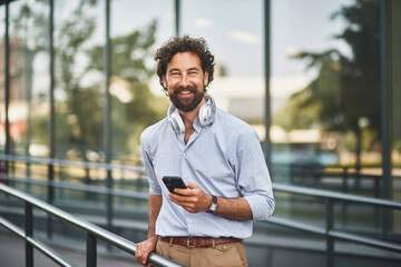 A smiling man stands casually with a smartphone in hand, wearing headphones, outside a modern...