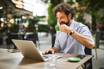 Man engaged in work on his laptop, smiling while sipping coffee at an outdoor cafe, surrounded by greenery and other patrons enjoying the pleasant weather.