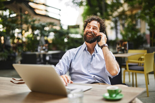 A man with curly hair and a beard chats on his phone while typing on his laptop at a cafe. The outdoor setting is vibrant with greenery, creating a relaxed work atmosphere.