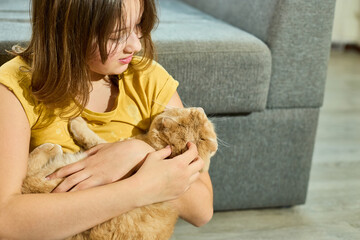 A teen girl relaxes at home, gently holding her Scottish Fold cat in her arms. They share a warm and affectionate moment on the living room floor, creating a serene atmosphere.