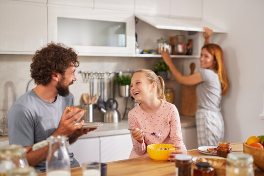 A father and daughter share a joyful moment over breakfast, with the daughter laughing while eating cereal. The mother is busy organizing items in the pantry, adding warmth to the morning.