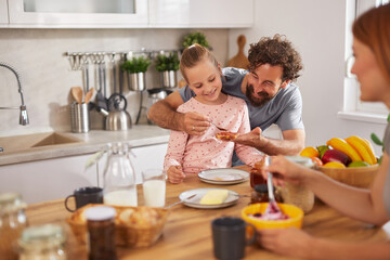 Family gathered in bright kitchen, savoring breakfast. Parents share laughter with daughter, spreading butter on toast and delighting in bright fruits and yogurt.