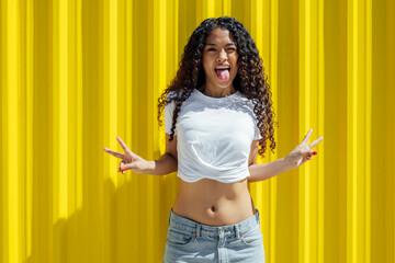 Woman gesturing peace sign with tongue out against yellow background