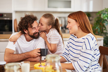 A father shares a loving moment with his daughter, who gives him a kiss, while the mother smiles nearby during breakfast in their cozy kitchen.