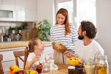 A cheerful family shares breakfast in a bright kitchen, with a mother bringing a basket of pastries...