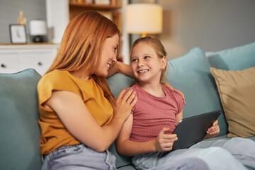 Obraz premium A mother and daughter share smiles while sitting together on a comfortable couch, exploring content on a tablet in their inviting living room filled with warm light.