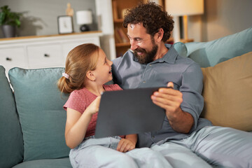 A father and daughter sit closely on a sofa, laughing and engaging with a tablet. They appear happy...