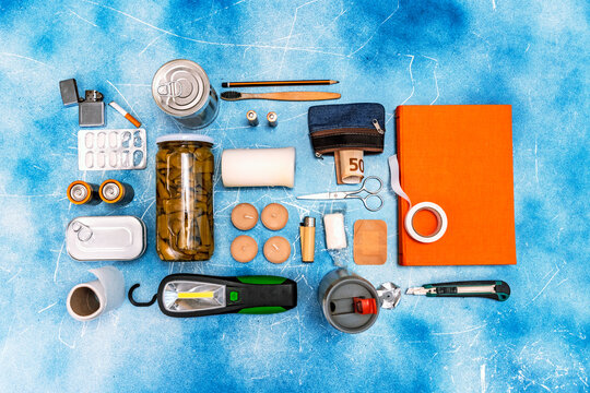 Flat lay of survival essentials kit with candles, lighter, batteries, and canned food on a blue background
