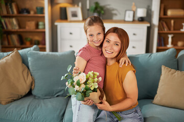 A young girl and her mother sit together on a comfortable couch, sharing smiles and joy. The girl holds a bouquet of flowers, creating a warm and loving atmosphere.