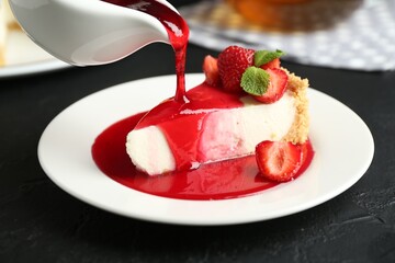 Pouring sauce onto piece of tasty cheesecake with strawberries and mint at black table, closeup