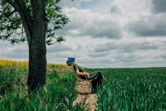 Woman sitting on a yellow folding chair under a tree in a field, reading a book with a cloudy sky above