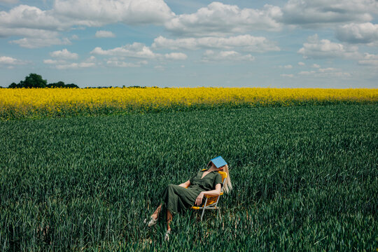 Woman relaxing on a yellow folding chair in a field of green wheat and yellow rapeseed, reading a book under a cloudy sky