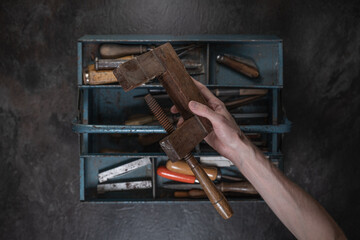 A hand holds a vintage wooden clamp above an open metal toolbox filled with old tools, photographed...
