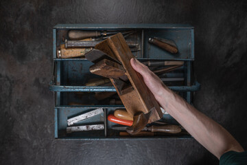 A hand holds a large vintage wooden hand plane above an open metal toolbox filled with old tools, photographed from above.
