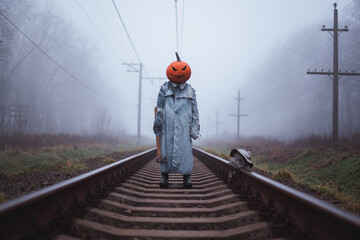 Terrifying person in carved pumpkin mask with axe and scarecrow severed head stands on misty...