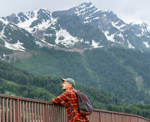 Young man hiker in a red plaid shirt and cap stands by a railing, gazing at majestic snow-capped mountains, surrounded by lush green forests, capturing a moment of tranquility in nature