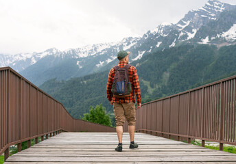 Man hiker in plaid shirt and backpack walking on wooden bridge surrounded by lush green mountains and snow-capped peaks, enjoying nature and outdoor adventure experience