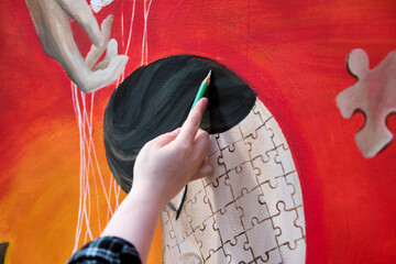 Close-up of adult female hand painting puzzle-themed mural. Vibrant red dominates, with canvas of...
