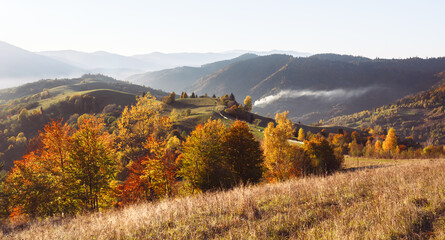 Rising sun lights up orange autumn trees on misty slopes beneath hazy peaks. Highland landscape in...