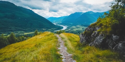 A scenic mountain trail winding through lush green hills with a river and blue mountains under a cloudy sky in the background.