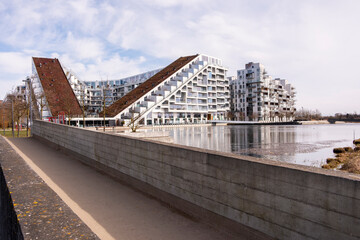 Modern waste-to-energy plant with dynamic sloping roof and urban riverside promenade under soft daylight in Copenhagen, featuring contemporary architecture and geometric design.