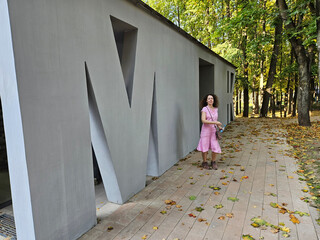 Woman standing in front of a building with the letter M on public toilet. She is wearing a pink...