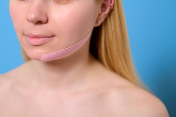 Obraz premium woman with facial lifting tape against blue background. close-up of innovative beauty trend for skin tightening and anti-aging. modern self-care and facial sculpting technique